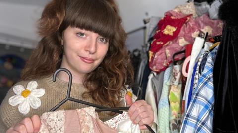 Amelie Rule, a young woman with a nose and septum piercing and thick brown wavy hair that has a blunt fringe and is partly tied up. She is wearing a brown knitted jumper with three-dimensional daisies on it and holds items of clothing on hangers, with a rack of clothing and fabric behind her on the right of the image