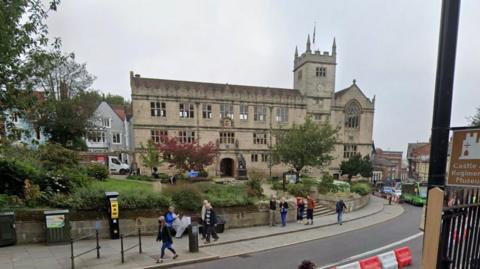 An old building with a statue of Charles Darwin sat in front of it. The building it sat back from the road, with people walking past on the path