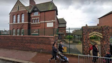 Red-brick primary school with a green metal fence and some parents meeting children at the school gates.