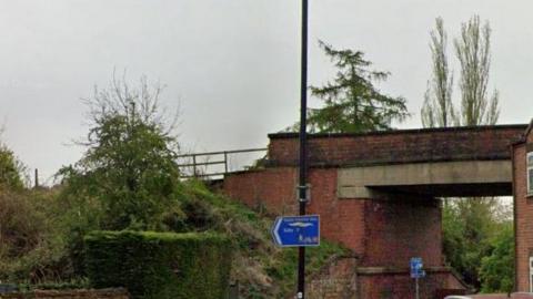 A roadbridge, pictured from the footpath beneath it. It is a brick structure and features a grass embankment to the side of it, as well as trees all around it.     