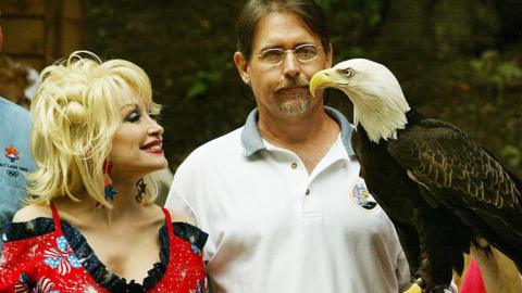 Dolly Parton, wearing an American-flag themed outfit, stands next to a man in a white polo shirt who is holding a bald eagle
