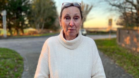 Chris Neen, a woman in a white jumper stand by a road outside her house. 