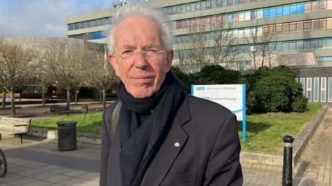 A man with white hair and glasses and wearing a black coat and scarf standing in front of a grey office building 