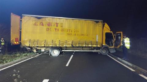 A yellow lorry is sideways and is across a road. Its back doors and open and the drivers side door is open. Two police officers are standing at either end of the vehicle.