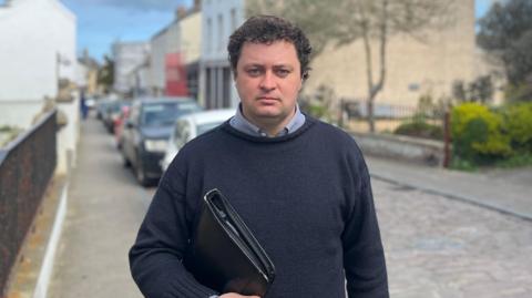 Alex Snowdon is wearing a navy knitted jumper over a light blue shirt as he stands on a cobbled street while carrying a large black folder.