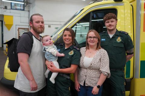 A group photos showing Dad Michael Everett who is wearing a grey t-shirt. Next to Michael is paramedic Courtney Mulligan who is wearing a paramedic uniform and holding baby Lydia Everett. Next to Courtney is mum Abie Everett wearing a blouse with black small spots on it. Next to Abie is another medic Corey Graham who is also wearing a medic's uniform and has his hands behind his back. All five are in front of an ambulance vehicle.