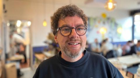 Tony Marks who has curly brown hair and beard, and glasses. Standing in his shop with customers sitting behind him and his chefs in the kitchen 