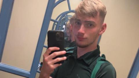 Jamie Kadolski poses for a selfie in a mirror. He is holding up a large phone with a black phone case. He is wearing a green polo neck and slightly smiling at the camera. He has short blonde hair and dark facial hair.