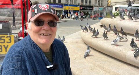 A pensioner with a navy jumper on and a red and black Gloucester Rugby cap smiles as he sits by his red mobility scooter in a city centre. He is sat next to some steps where pigeons have gathered and a row of shops can be seen behind him.