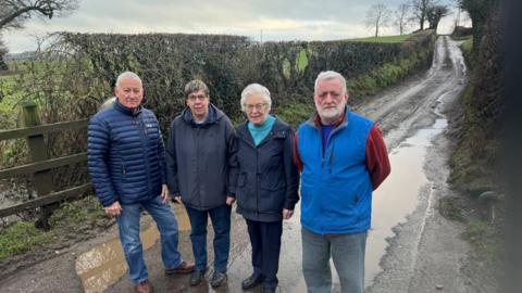 Two men (left and right) and two women (centre) standing in a rural lane which runs up a hill behind behind them. Water can be seen running down the hill and pooling in damaged parts of the road near where the group is standing.