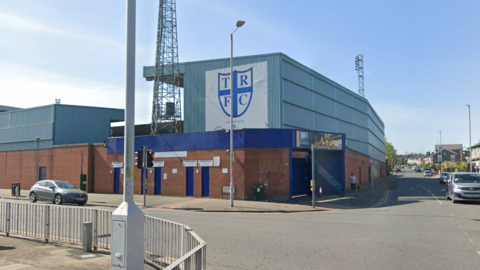 Google Streetview image of Tranmere Rovers' Prenton Park stadium. Taken from outside the ground, two large stands are visible. One of them includes a large blue-and-white shield with the initials TRFC printed on it.