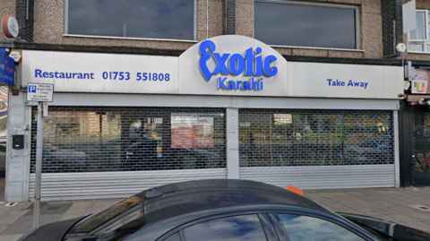A Google Maps picture of Exotic Sweet Centre, which has a silver and blue frontage with shutters pulled down. It also says restaurant and take away, with a phone number on the front of it too.