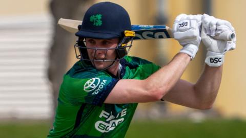 Lorcan Tucker, in dark blue helmet and mainly green cricket shirt, watches the ball after playing a shot, with the bat over his left shoulder as he looks to his right