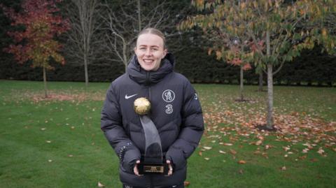 Hannah Hampton is holding the trophy which has a black base and small golden football at the top. She is standing in a grassy area with trees in the background, and is wearing a black padded Chelsea branded coat. 