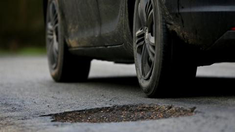 A black car parked with its rear wheel next to a large pothole in the road surface.