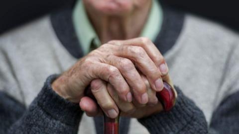 An older man, wearing a grey jumper, resting his hands on a red walking stick.