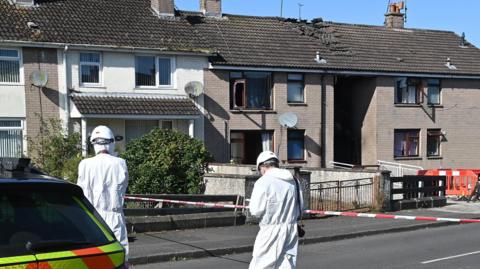 Two forensics pictured next to a police car and in front of the houses where the fire occurred. Red and white tape is in place to barricade the area. The roof of one of the houses is visibly damaged and the windows have been blown out.