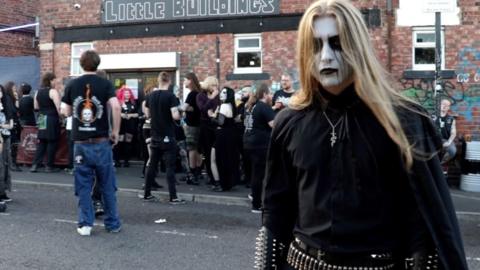 Envus is stood in front of a crowd outdoors. He is wearing all black, has long brown hair and his face painted white with black accents.