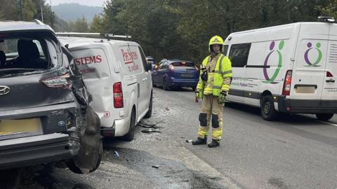 A wide image of two damaged vehicles on the road. There are bits of debris on the floor. A fire officer is stood in-between the parked damaged cars and the other vehicles driving on the road. 