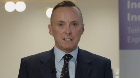 Tom Crowther wearing a black suit, blue shirt and black printed tie looking at the camera