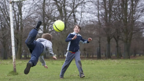 Two boys play football on a field. One boy is stood in a goal while the other is captured in the air kicking the ball over his head from behind. 