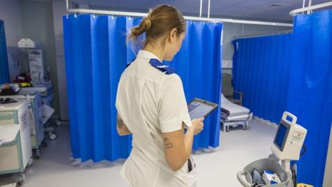 A maternity unit ward with a medic standing in the centre, facing away from the camera and holding a tablet device. They wear a light-coloured uniform with blue trim and has hair tied back. Blue privacy curtains hang in sections around examination bays, partially enclosing a hospital bed visible in the background. To the right, a medical trolley holds monitoring equipment and devices with screens and cables. On the left, additional hospital equipment and storage units line the wall.