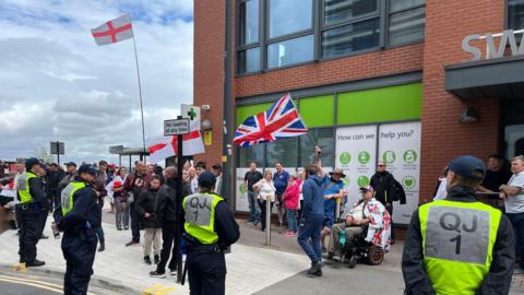 A group of people stand outside a green-panelled building holding Union Jacks and England flags. the are four people marshalling wearing high-viz jackets