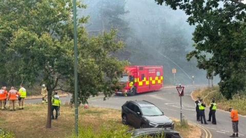 Emergency service workers in hi-vis jackets congregate in two groups - one group of police and one group of firefighters, in the road next to a fire engine. There are two parked cars between the group. Thick smoke is visible between the trees.