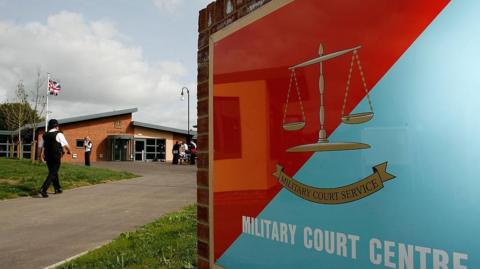 A view of the Military Court Centre in Bulford. There is a red and blue sign at the forefront of the picture with the building in the background. 