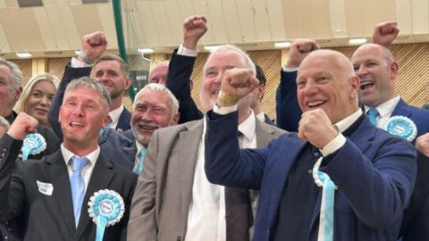 A crowd of Reform party members wearing rosettes. They are wearing suits and cheering.