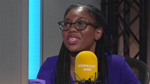 Kemi Badeonoch speaking on Newscast, Her hair is in braids, she is wearing glasses a purple top and is speaking into a yellow mic with Newscast 91ȱ printed 