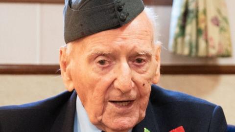 A World War Two veteran wearing a blue jacket and miiltary beret. He is looking directly into the camera with his mouth slightly open.