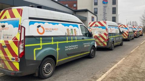 Five Wales & West Utilities vans lined up in a queue at the site of the incident. They are parked outside a Travelodge, near a construction site. 