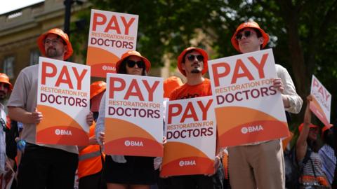 Three young men and a woman stand in a line at a doctors' strike. They are all wearing identical orange bucket hats and holding placards towards the camera which say 'pay restoration for doctors'. A tree and a crowd of protesters are visible behind them.