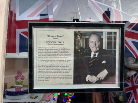 A framed image of a man in a suit and tie next to a text of Enoch Powell's "Rivers of Blood" speech. There are Union flags in the background.