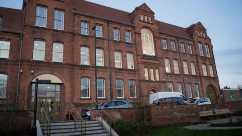 A grand building. It has lots of windows and stands tall. It has a modern entrance and there are some cars parked outside.