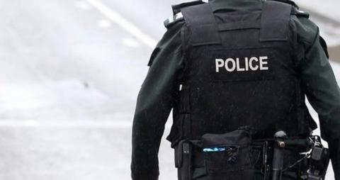 A police officer is standing on a road. They are in a black uniform and have a belt with a number of items. 
