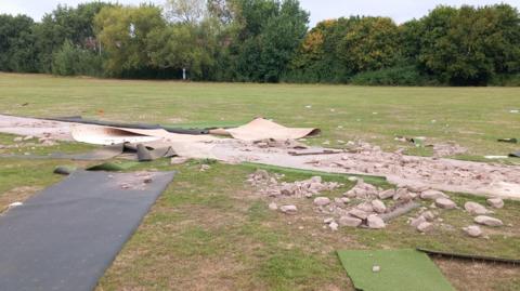 The cricket pitch is badly damaged, with ripped artificial turf and debris scattered all over the field. Behind it, trees and a few buildings can be seen in the distance.