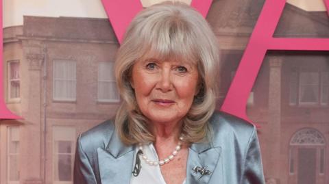 Dame Jilly Cooper smiles as she wears a silk, pale blue blazer, at a Rivals red carpet event. She is also wearing a pearl necklace and a silver pin of someone riding a horse.