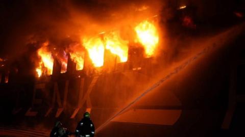 A ship is seen on fire and first responders are seen trying to extinguish the flames.