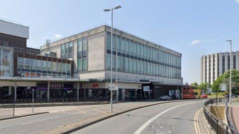 A large brick and concrete panel building with windows on its top floor - people can be seen walking past it on the pavement.