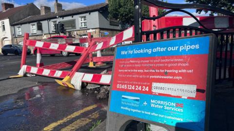 A water leak on a busy main road which has been cordoned off with a Yorkshire Water sign stating We're looking after our pipes