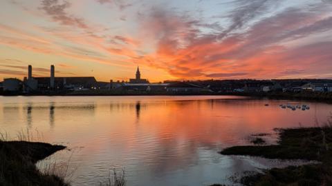 A vivid sunset casts orange and pink reflections across a calm body of water, with a town skyline— including tall chimneys and a central tower— silhouetted against the sky.