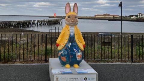A large Peter Rabbit sculpture dressed in a yellow fisherman's hat and jacket standing in front of the sea. Underneath it is wearing blue dungarees with various fishing equipment painted on. There is a black fence behind it with some shrubbery behind it, before the sea. There are buildings on the right and a small red lighthouse immediately behind it on a pier.