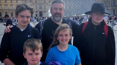 Jonathan Sivyer is standing in the middle of his four children in front of the iconic Louvre Museum pyramid.