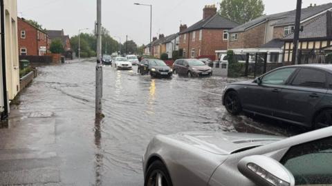 A road is flooded and several cars are driving through it on either side of the road. The water has come up on to the pavement is going near people's houses either side of the road. 