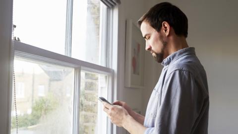 A person looking down at a phone. They are standing side on so we can see their profile from the waist upwards. They are inside facing a large window. 