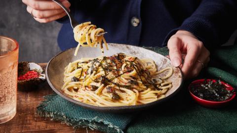 A person holding a spoonful of linguine pasta from a circular bowl, on green cloth