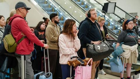 Various people wait, looking a bit bored, at an airport with their luggage.