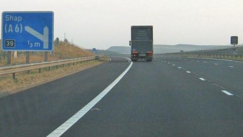 The M6 road approaching junction 39 at Shap. There is a blue motorway sign showing the direction to Shap. There is a lorry travelling on the motorway.